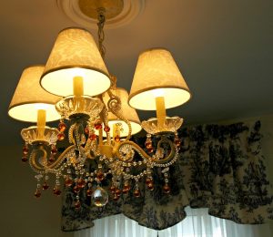 French Country Style Chandelier: A small light fixture with four lights and four shades on top of those lights.